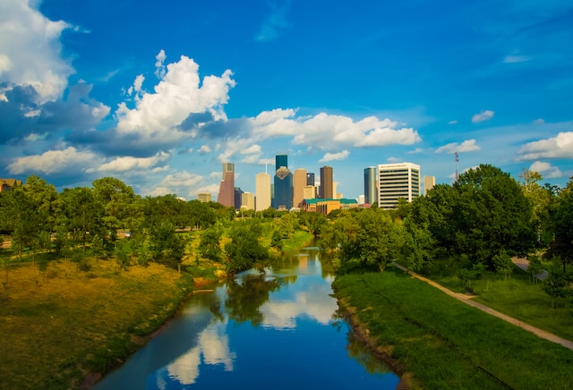 A view of a stream surrounded by green grass and trees in Houston, Texas, with some skyscrapers